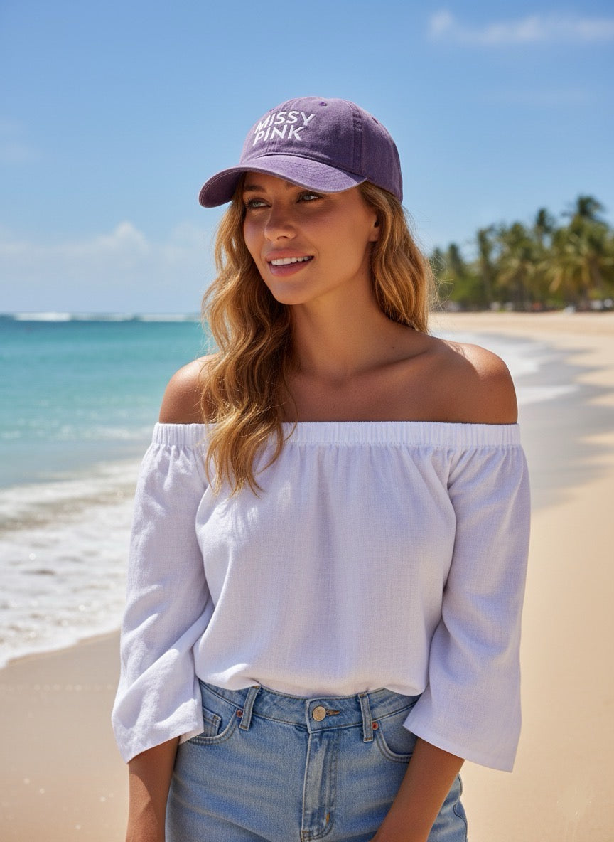 Woman wearing a purple missy pink cap and white off-shoulder top on a beach